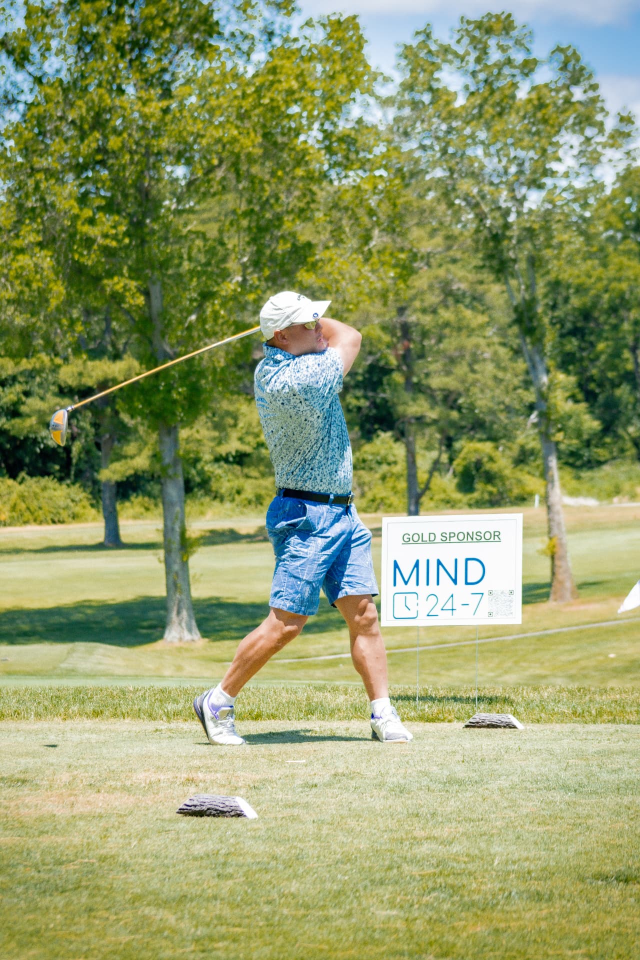 Golfer finishing a tee shot beside a gold sponsor sign